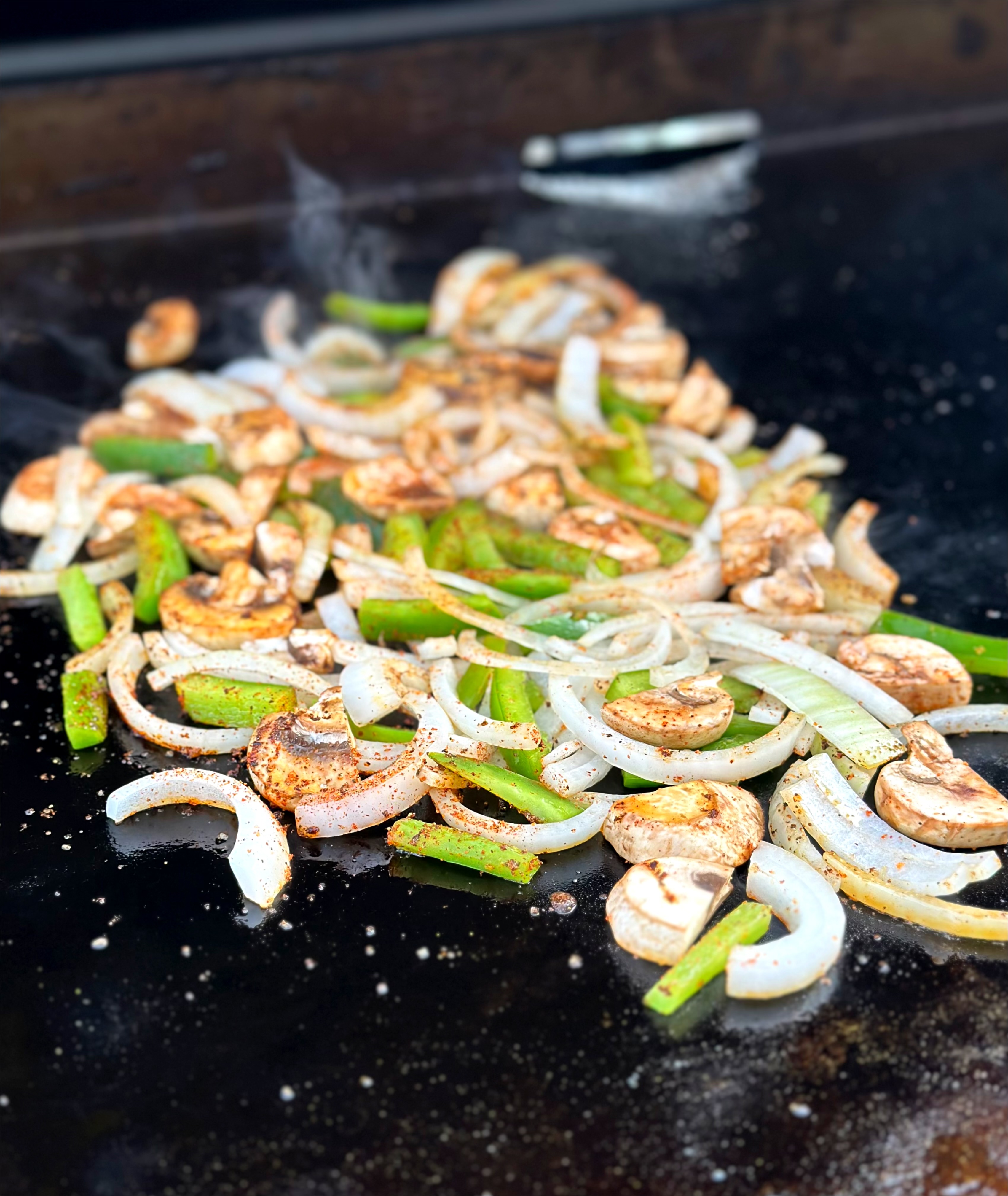 Onions, bell peppers, and mushrooms seasoned and cooking on the griddle. 